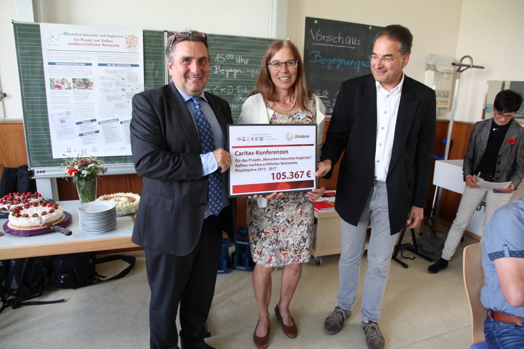 Zwei Männer überreichen einer Frau einen Scheck über 105.367 € in einem Klassenzimmer.