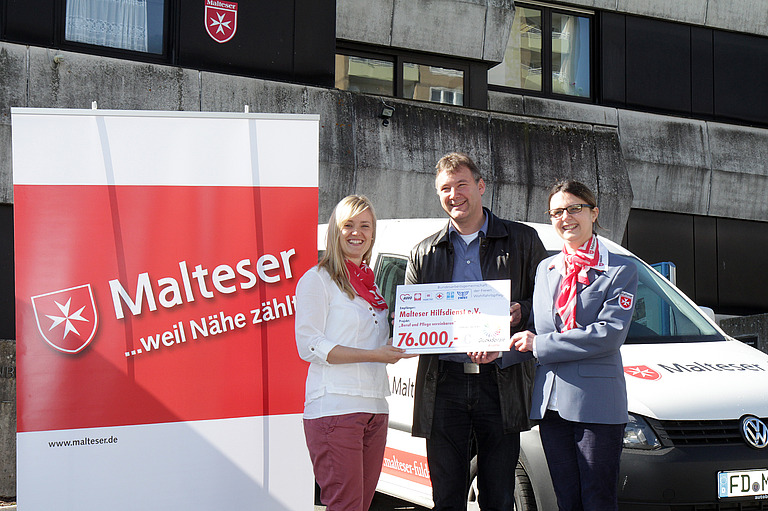 Drei Personen stehen vor einem Malteser-Rollup und halten einen Scheck über 76.000 € in die Höhe.