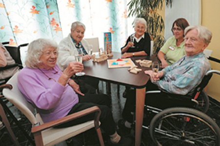 Fünf ältere Frauen, die gemeinsam an einem Tisch sitzen, ein Spiel spielen und Sekt trinken.