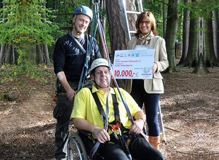 Ein Rollstuhlfahrer und ein Mann stehen in Kletterausrüstung in einem Kletterpark. Eine Frau hält einen Scheck über 10.000 €.